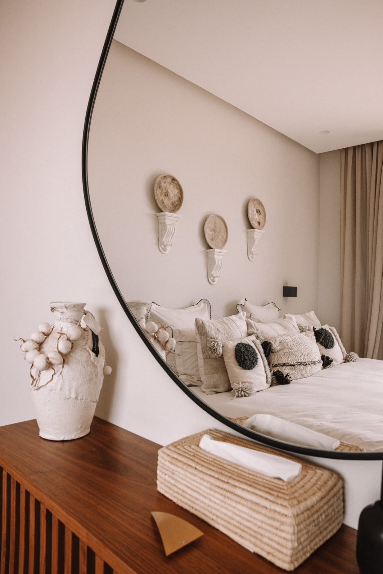 Bedroom reflected in a large round mirror above a wooden sideboard, with woven basket and ceramic vase